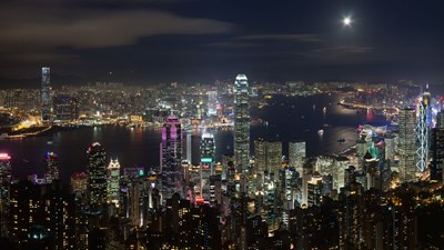A panoramic view of Hong Kong at night, with the skyscrapers of  Central lit up against the dark sky. The Victoria Harbour is also visible, with the Star Ferry crossing in the foreground.