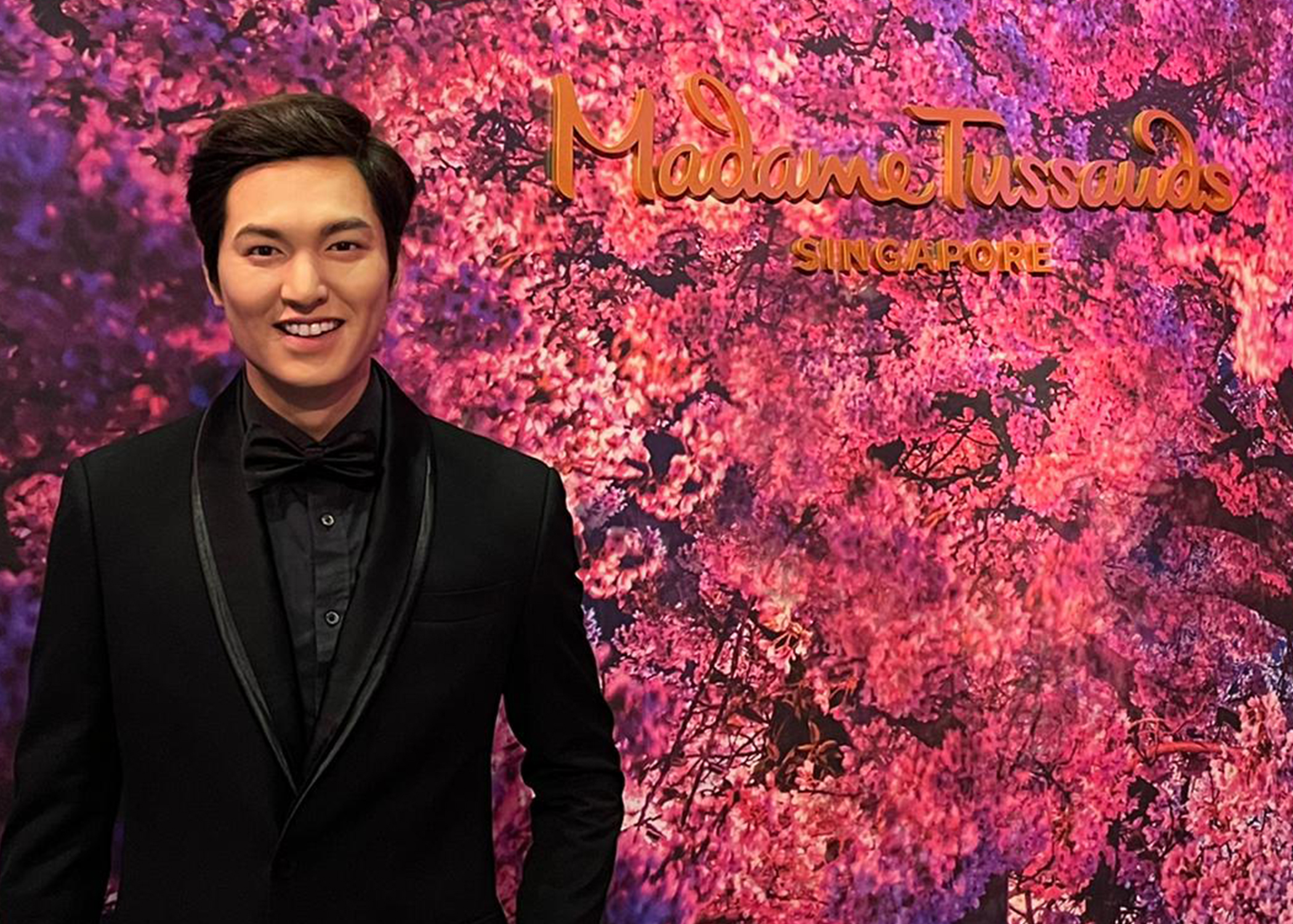 At Madame Tussauds Singapore, the wax figure of Korean celebrity and actor Lee Min Ho, dressed in a black suit and ribbon tie, smiles while standing against a backdrop of cherry blossoms