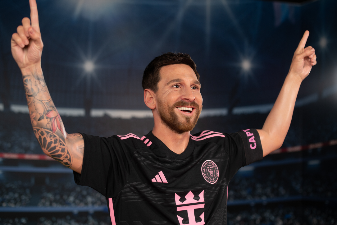 A wax figure of Lionel Messi at Madame Tussauds Orlando, dressed in an Inter Miami CF jersey, smiling with both arms raised triumphantly in a stadium setting illuminated by bright lights.