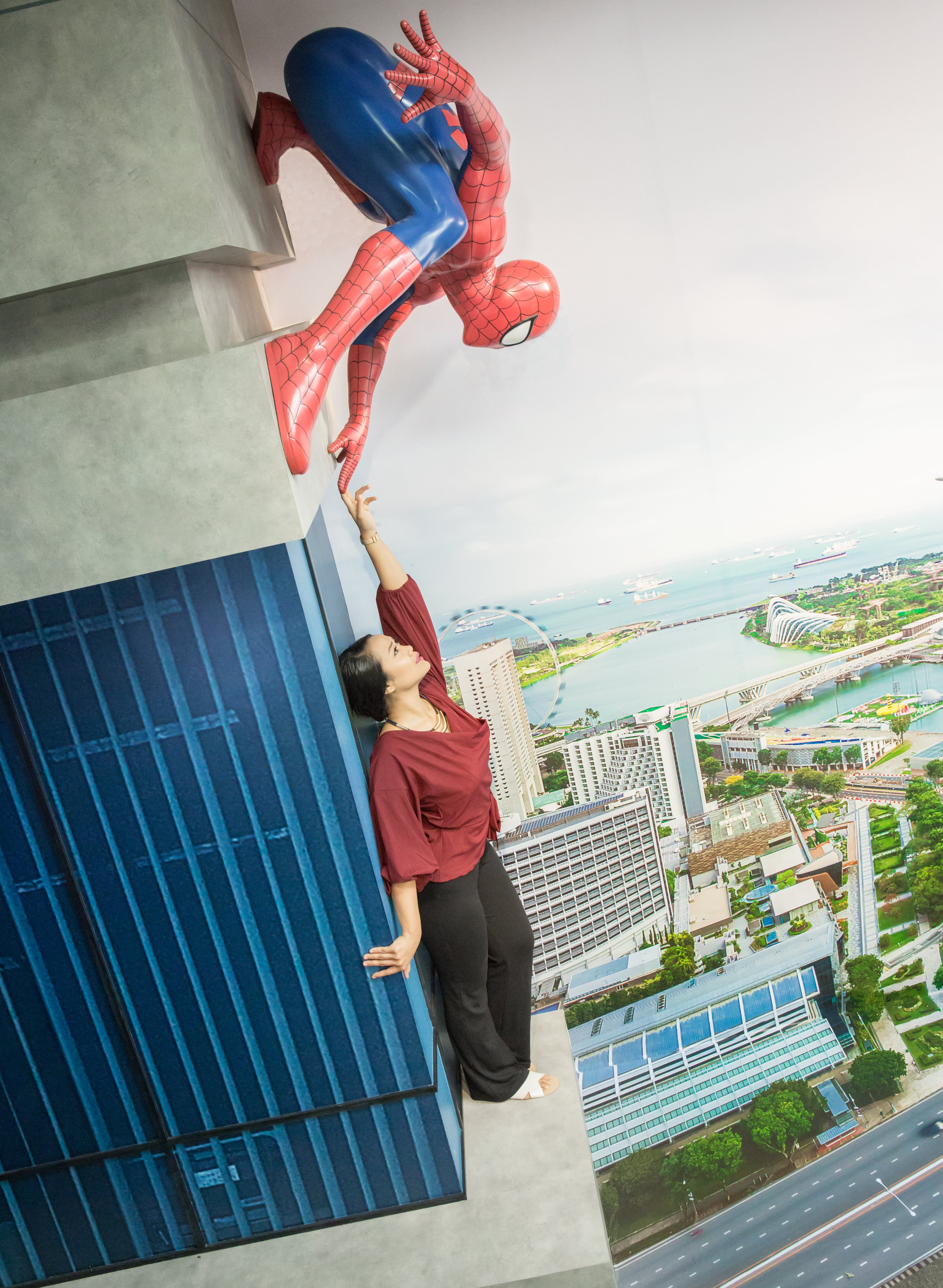 A visitor at Madame Tussauds Singapore - Marvel Universe 4D, pointing towards a Spider-Man figure that appears to be clinging to the side of a building exterior. The visitor is standing in front of a cityscape backdrop, making it seem like they are part of a thrilling superhero adventure