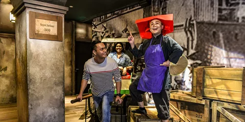 A set up of Singapore's history in the 1900s, depicting Keppel Harbour. A realistic wax figure of a Samsui woman beams as she stands on two boxes. A happy man holds the handles of the rickshaw and acts like he is pulling it with a girl seated behind.
