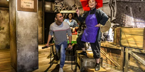 A set up of Singapore's history in the 1900s, depicting Keppel Harbour. A realistic wax figure of a Samsui woman beams as she stands on two boxes. A happy man holds the handles of the rickshaw and acts like he is pulling it with a girl seated behind.