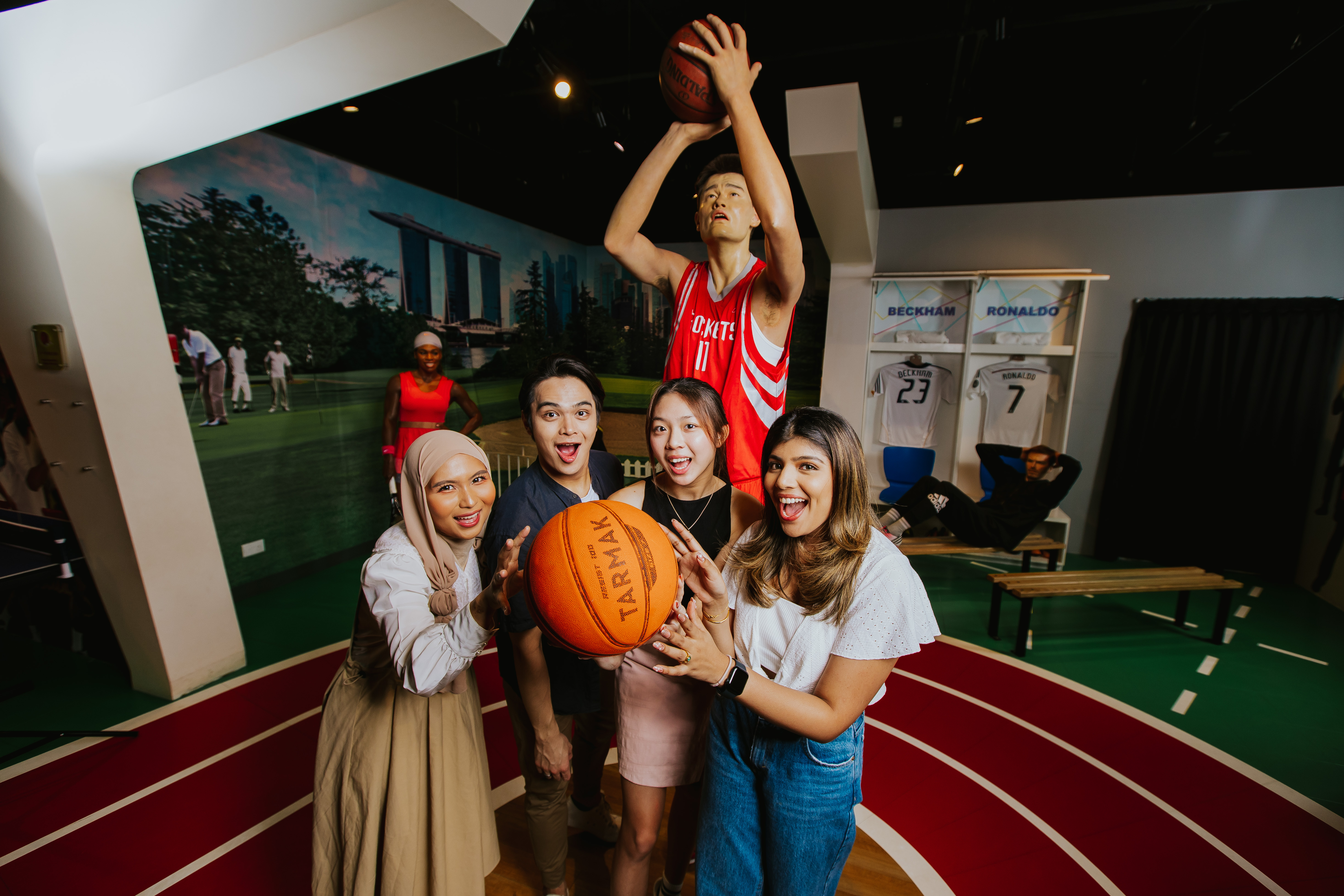 At Madame Tussauds, three girls and a boy happily mimic holding a basketball in front of Yao Ming's wax figure. His impressive height, emphasized by shooting a basketball in the background, highlights his title as the tallest Chinese NBA player. 